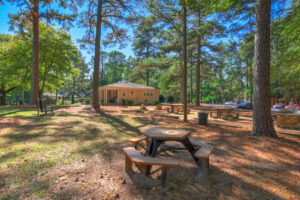 Exterior Picnic area, leasing office in background, multiple picnic tables, outdoor rocking chair, lush foliage surrounding the area, photo taken on a sunny day.