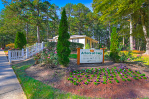 Exterior leasing office, arbors at cary signage, fresh flowers planted around signage, meticulous landscaping, lush foliage in background, photo taken on a sunny day.