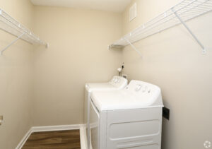 Interior laundry room, white washer and dryer, 2 shelves, neutral toned walls, wood floors.